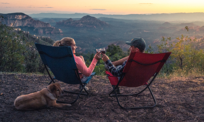 A man and a woman sitting in lawn chairs clinking glasses while looking out over a beautiful canyon.