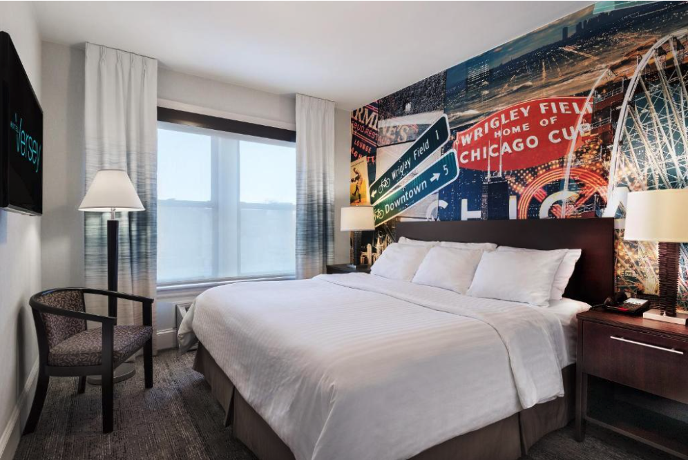A hotel room with a double bed, a chair, and lamps, with a mural of Chicago landmarks on the wall behind the bed.